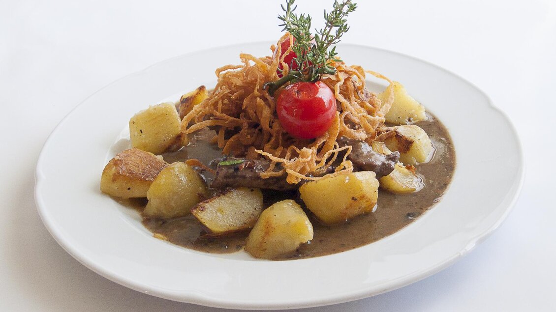Ein teller mit zartem Fleisch, knusprigen Zwiebelringen und goldenen Kartoffeln. Oben dekoriert mit einer Kirschtomate und frischen Kräutern. | © Tscheppe
