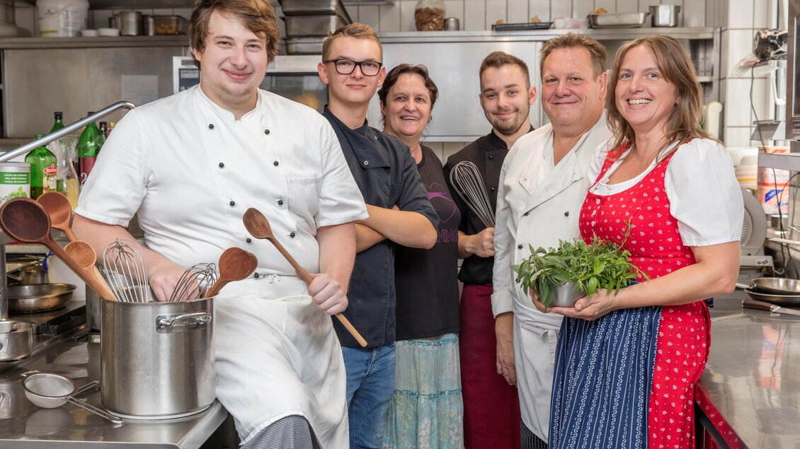Eine fröhliche Küchenteam, das in einer Restaurantküche steht. Sie halten Kochutensilien und frische Kräuter in der Hand. | © Tscheppe
