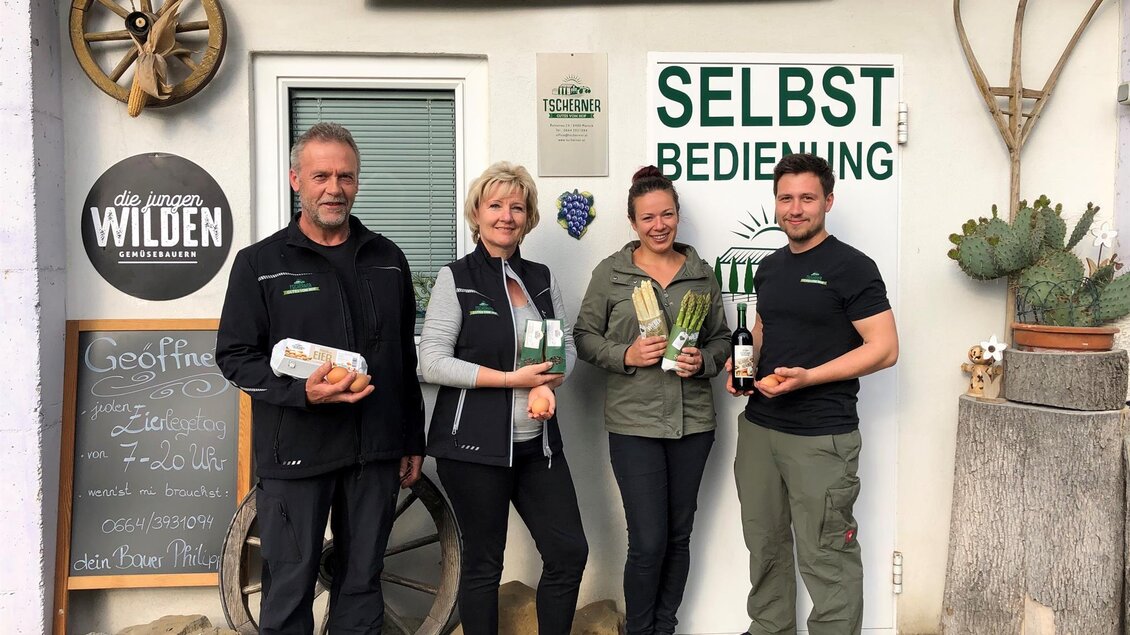 Eine Gruppe von vier Personen steht vor einem Bauernladen. Sie halten frische Produkte in den Händen und zeigen ein einladendes Lächeln.