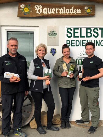 Eine Gruppe von vier Personen steht vor einem Bauernladen. Sie halten frische Produkte in den Händen und zeigen ein einladendes Lächeln.