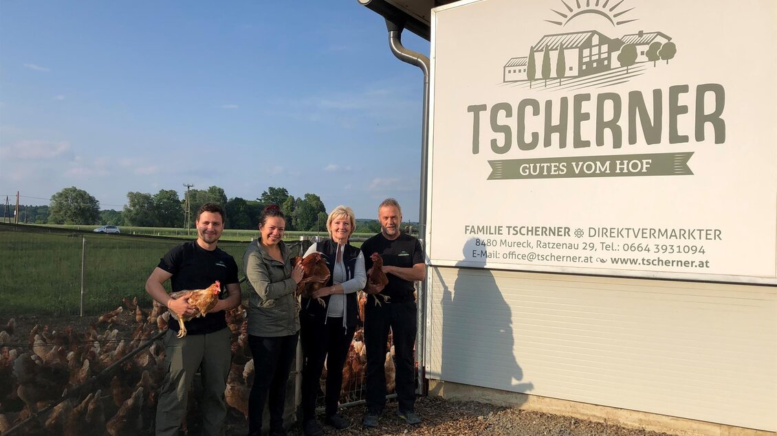 Eine Gruppe von vier Personen steht vor einem Bauernhaus mit einem Schild, das "Tscherners Gut vom Hof" zeigt. Sie halten jeweils ein Tier in den Armen und lächeln freundlich.