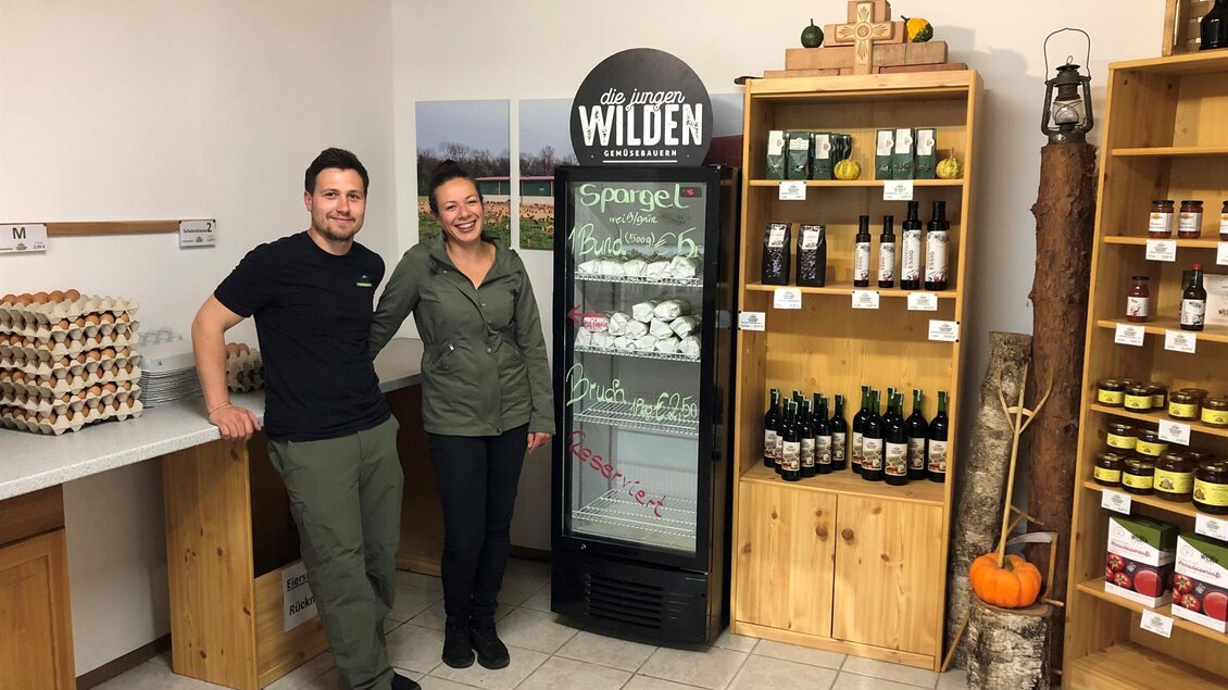 Zwei Personen stehen in einem kleinen Geschäft mit Holzmöbeln. Im Hintergrund sind Produkte wie Getränke und Eier zu sehen.