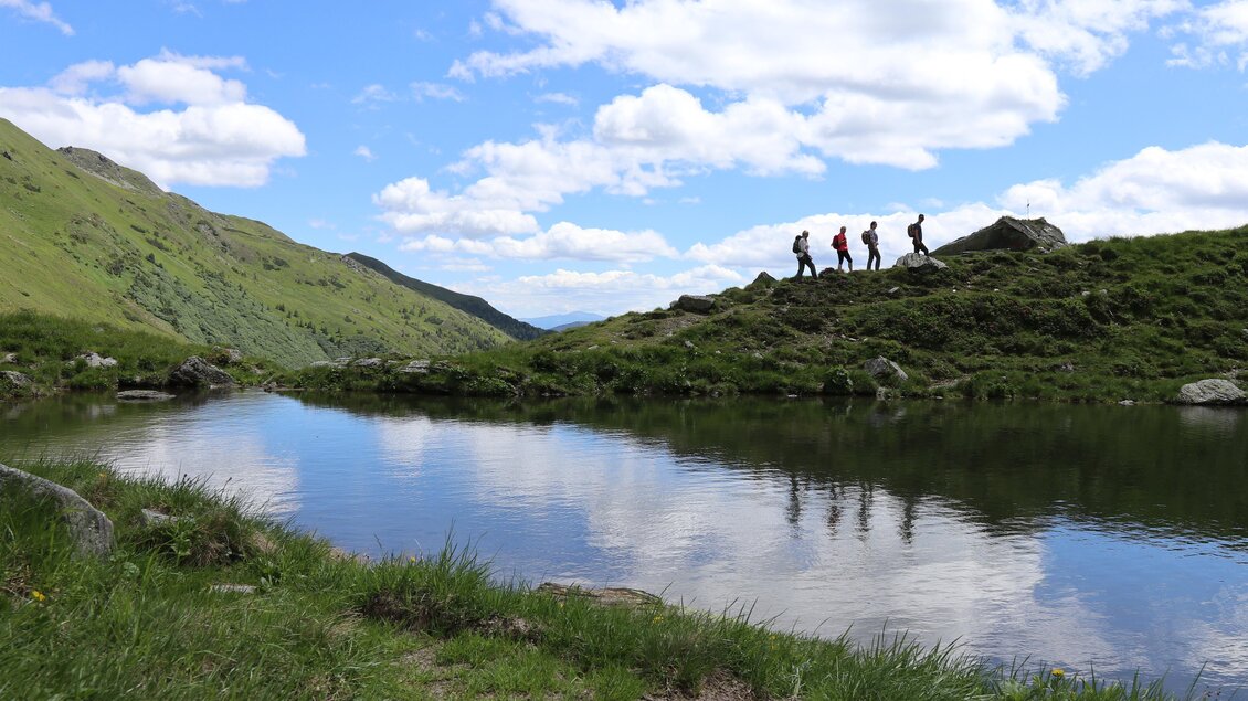 Eine Gruppe von Wanderern steht auf einem Hügel neben einem ruhigen See. Der Himmel ist blau mit weißen Wolken und die Umgebung ist mit grünem Gras und Bergen geprägt. | © Weges
