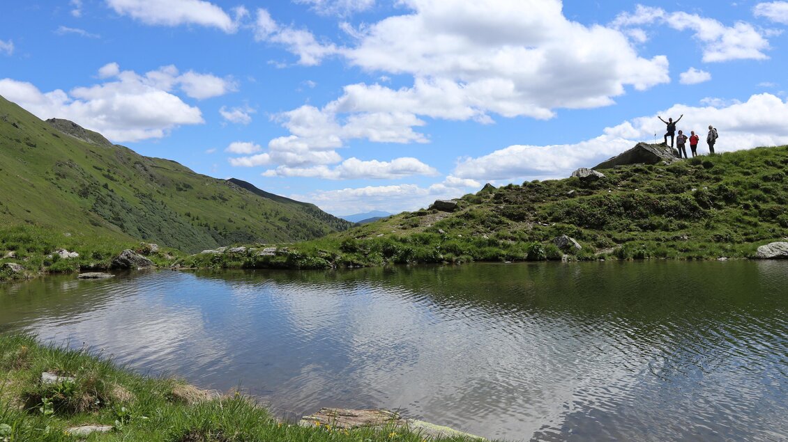 Ein idyllischer Bergsee mit grünem Ufer und sanften Hügeln im Hintergrund. Im Hintergrund sind Personen, die die Aussicht genießen. | © Weges