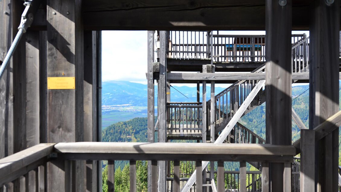 Eine Holzstruktur mit mehreren Ebenen und Treppen. Im Hintergrund sind grüne Berge und ein klarer Himmel zu sehen. | © Anita Fössl