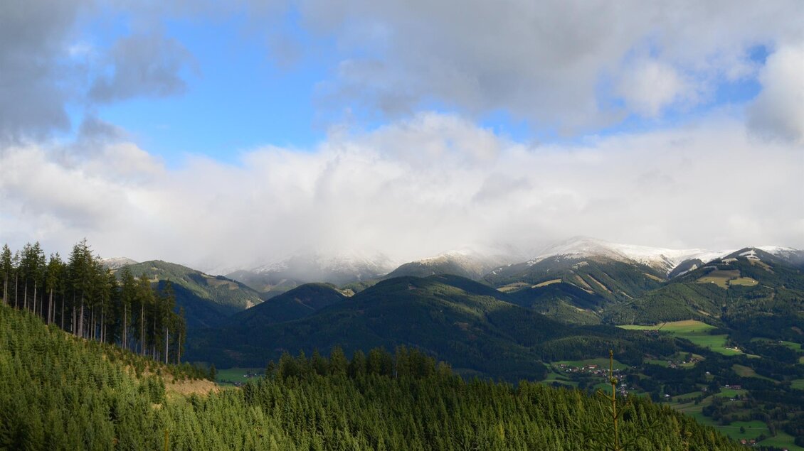 Eine malerische Berglandschaft mit grünen Wäldern und schneebedeckten Gipfeln. Der Himmel ist teilweise bewölkt und sorgt für eine stimmungsvolle Atmosphäre. | © Anita Fössl
