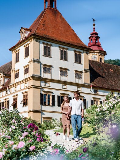 A couple walks along the gravel path in the castle park, with Schloss Eggenberg in Graz visible behind them. | © Graz Tourismus-Mias Photoart