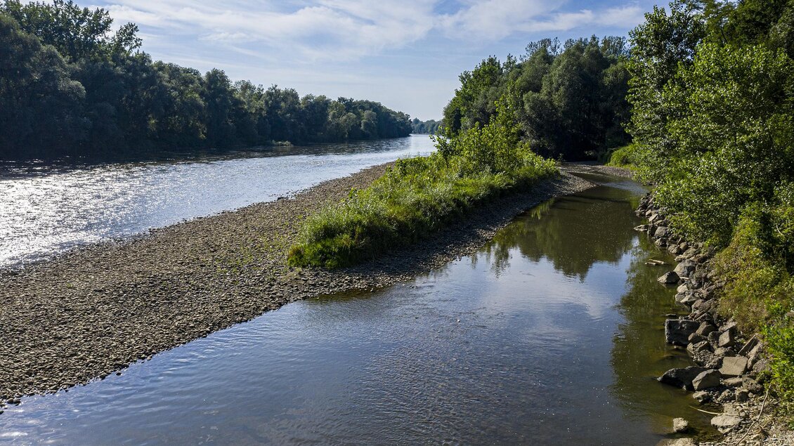 Ein ruhiger Fluss mit klarem Wasser und grünem Uferbewuchs. Die Landschaft ist von Bäumen und strahlendem Himmel umgeben. | © Thermen- & Vulkanland