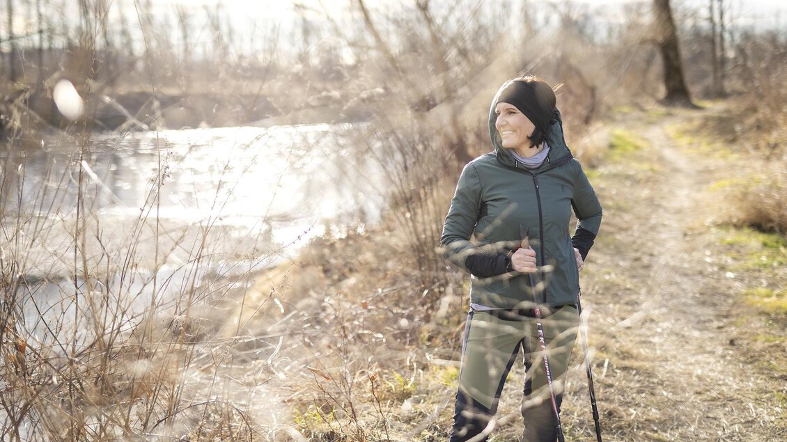 Eine Frau steht auf einem Wanderweg neben einem Wasserlauf. Sie trägt sportliche Kleidung und blickt in die Ferne. | © Thermen- & Vulkanland