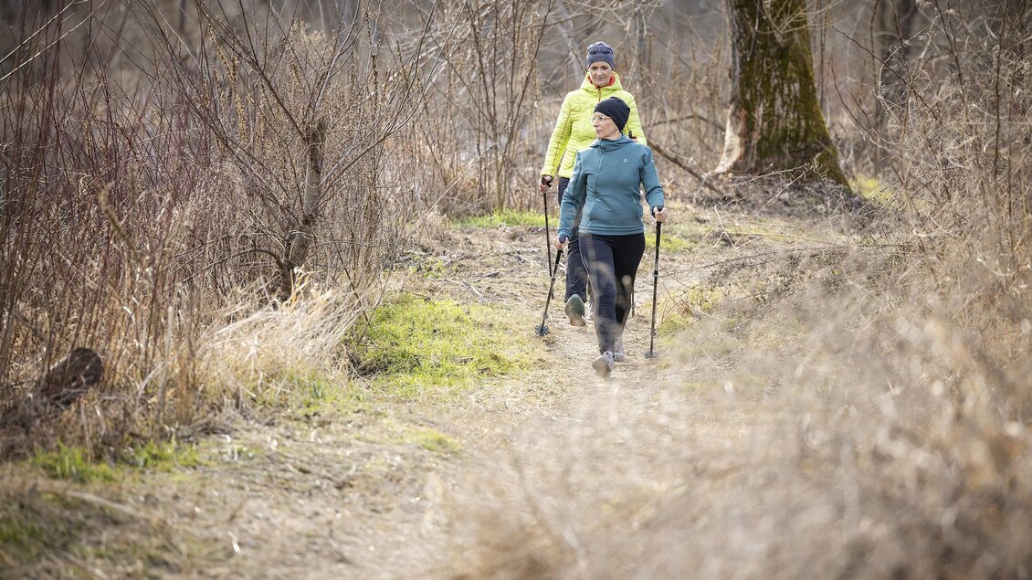 Zwei Personen wandern auf einem schmalen Weg durch die Natur. Sie tragen warme Kleidung und nutzen Wanderstöcke. | © Thermen- & Vulkanland