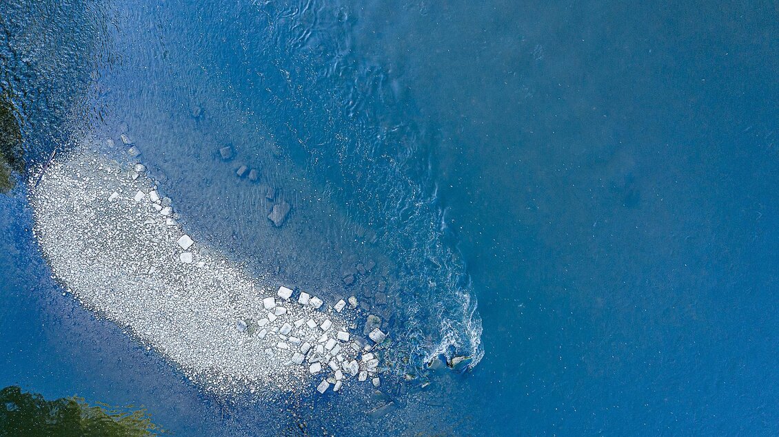 Ein ruhiger Gewässerabschnitt mit einer kleinen Kiesinsel und einigen Steinen. Das Wasser hat eine klare, blaue Farbe. | © Thermen- & Vulkanland