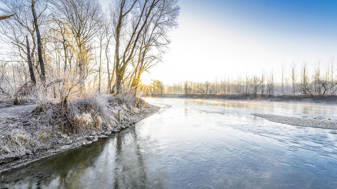 Eine ruhige Flusslandschaft bei Sonnenaufgang, umgeben von frostigen Bäumen. Das Wasser spiegelt den klaren Himmel wider. | © Thermen- & Vulkanland