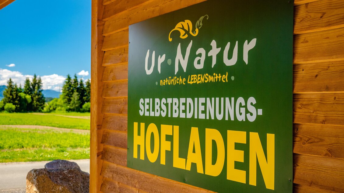 Ein Schild des Hofladens "Ur-Natur" mit den Worten "Selbstbedienungs-Hofladen". Im Hintergrund sind grüne Wiesen und blaue Himmel sichtbar. | © Wolfgang Speckner