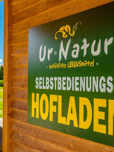 A sign of the farm shop "Ur-Natur" with the words "Self-Service Farm Shop". In the background, green meadows and blue skies are visible. | © Wolfgang Speckner