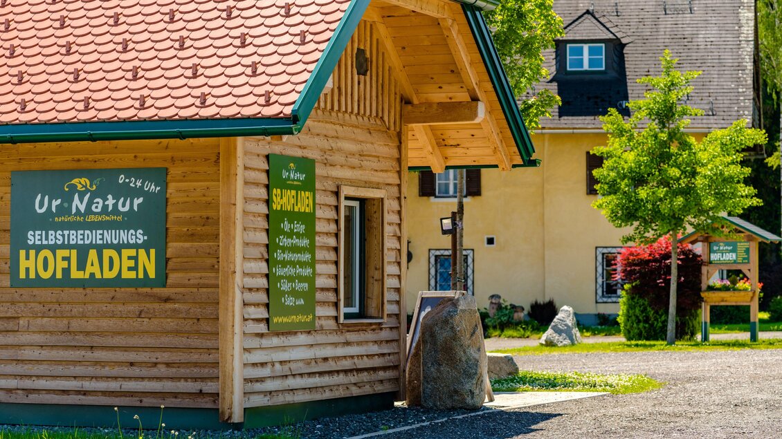 Ein kleiner Holzladen mit einem roten Dach steht in einem ländlichen Gebiet. Umgeben von grünen Bäumen und einem klaren Himmel, bietet der Laden regionale Erzeugnisse an. | © Wolfgang Speckner