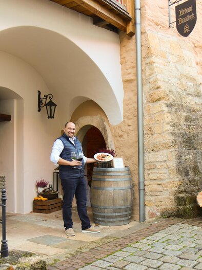 A friendly waiter stands in front of a historic building holding a dish. The surroundings are inviting with a wooden barrel and a beautiful garden. | © Winkler-Hermaden