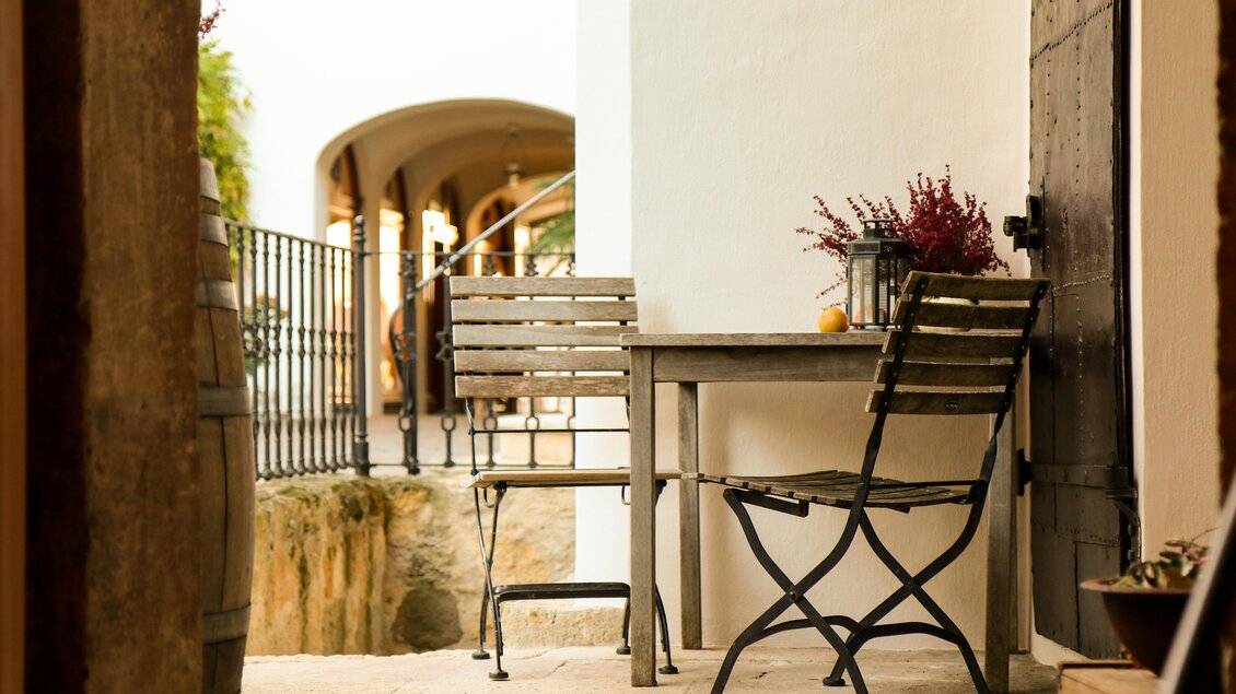 Ein schöner Außenbereich mit einem Holztisch und zwei Stühlen. Im Hintergrund sind Pflanzen und eine architektonische Struktur sichtbar. | © Winkler-Hermaden