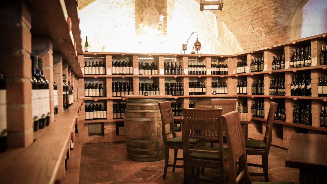 Ein gemütlicher Weinkeller mit Regalen voller Weinflaschen. In der Mitte stehen Holzstühle und ein Fass. | © Winkler-Hermaden