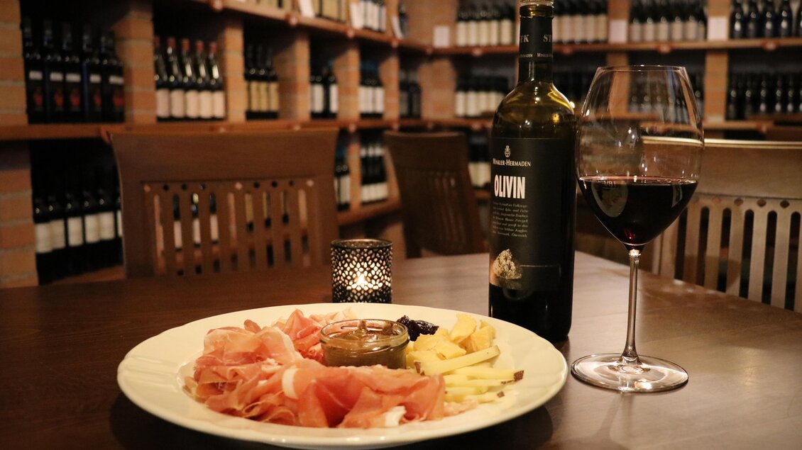 Ein Teller mit Schinken, Käse und Oliven, begleitet von einer Flasche Rotwein und einem Glas. Im Hintergrund sind Regale mit Weinflaschen sichtbar. | © Winkler-Hermaden