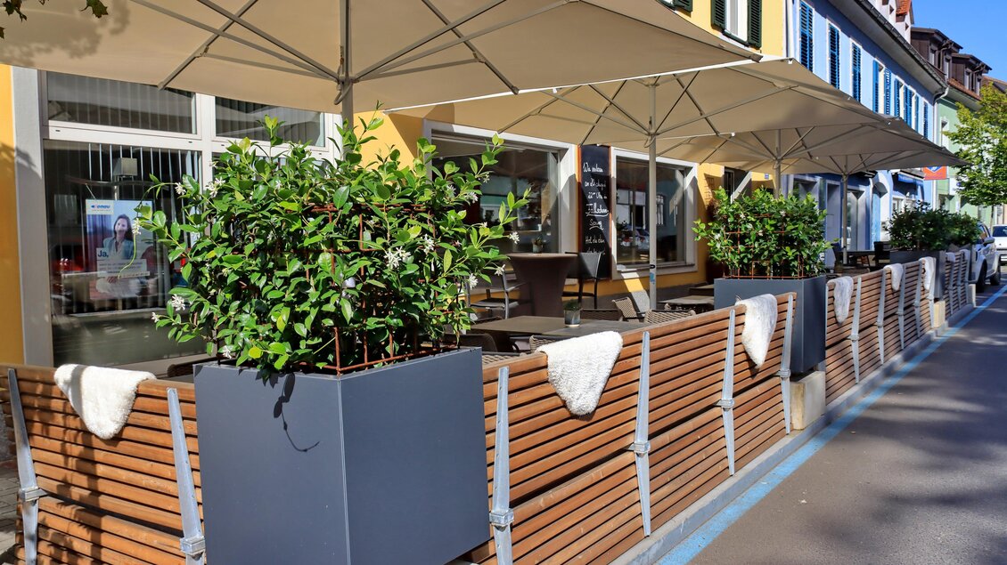 Ein gemütlicher Außenbereich eines Cafés mit Sonnenschirmen und Pflanzen. Die Holzwände und Blumenkübel verleihen dem Platz eine einladende Atmosphäre. | © © Urbanistub'n