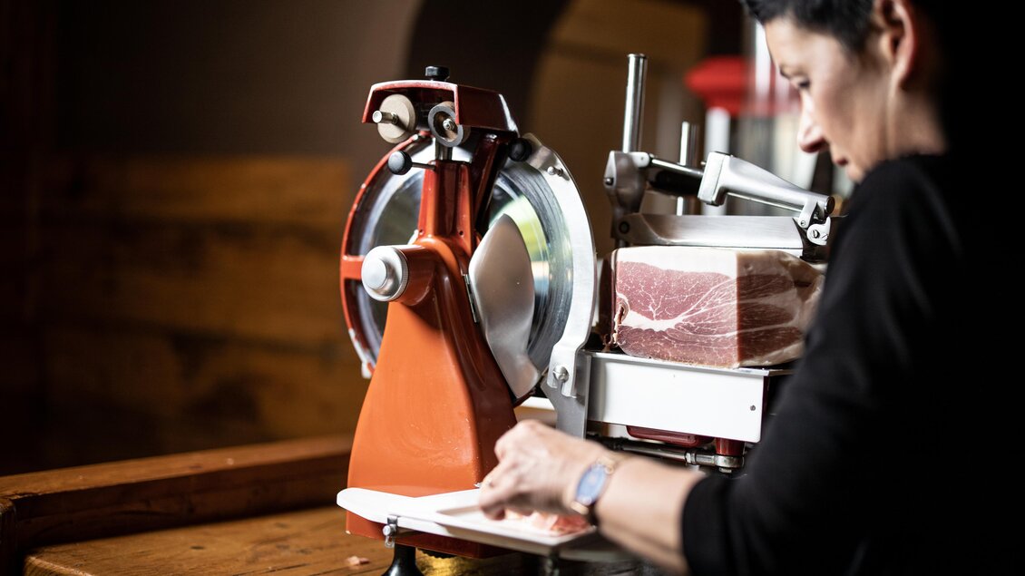 Eine Person schneidet mit einer Aufschnittmaschine sorgfältig Schinken. Die Umgebung wirkt rustikal und einladend. | © © Urbanistub'n