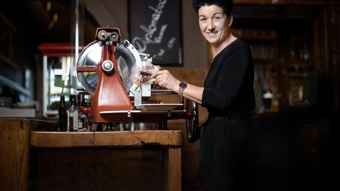 Eine Frau steht an einer Wurstschneidemaschine in einem gemütlichen Restaurant. Sie lächelt freundlich und scheint gerade etwas zuzubereiten. | © © Urbanistub'n
