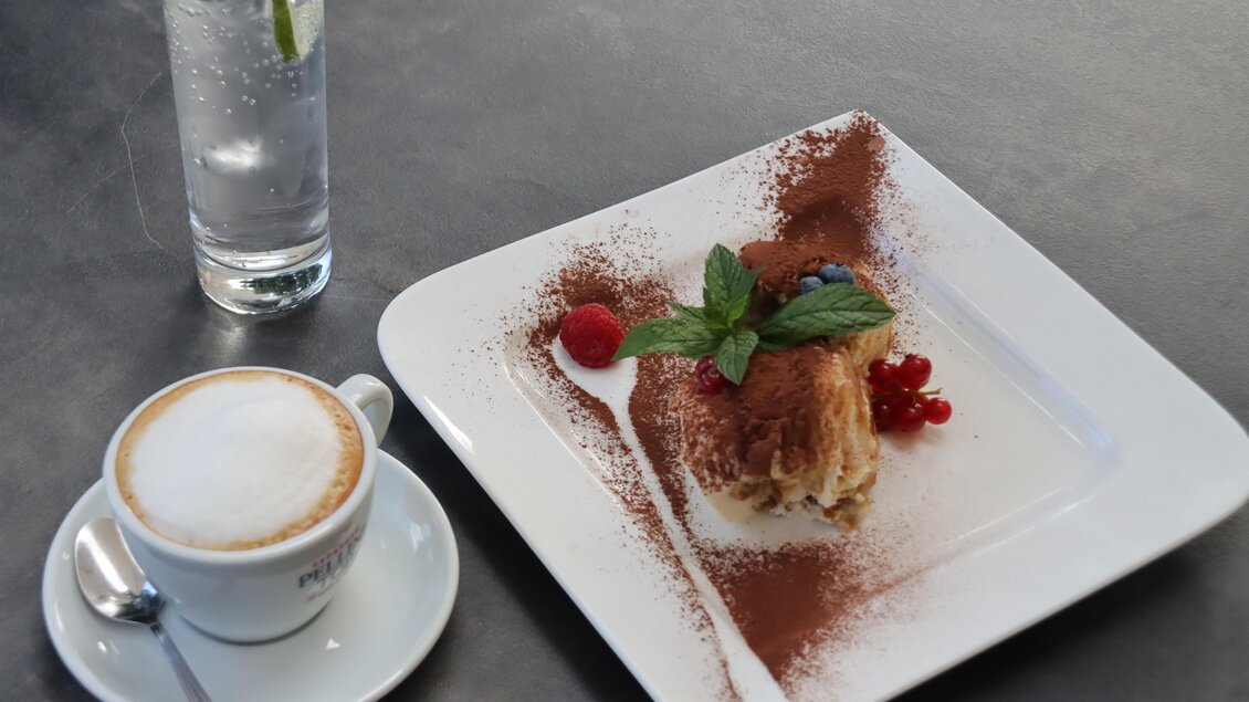 Ein Teller mit Dessert, dekoriert mit Beeren und Minze, liegt auf einem Tisch. Daneben steht ein Glas Wasser mit einer Limettenscheibe und eine Tasse Kaffee. | © © Urbanistub'n