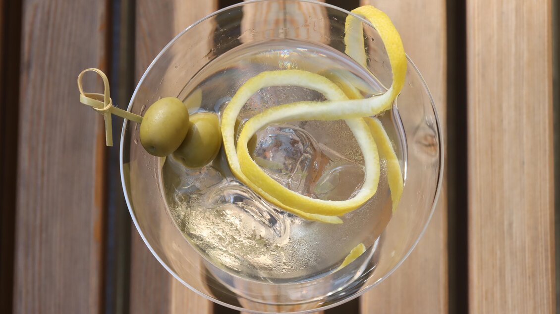 Ein Cocktailglas mit eisgekühltem Getränk, einer Olive und einer Zitronenscheibe dekoriert. Der Hintergrund zeigt eine Holzoberfläche. | © © Urbanistub'n