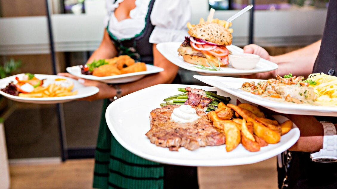 Ein Kellner serviert verschiedene Gerichte auf Tellern, darunter ein Steak und ein Burger. Im Hintergrund sind weitere Speisen sichtbar. | © Urdlwirt Hotel - Restaurant