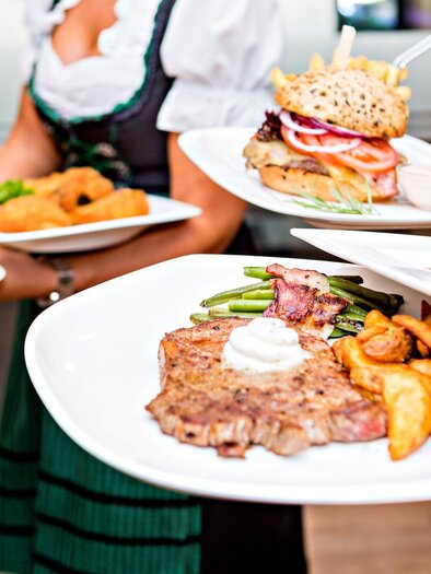 A waiter serves various dishes on plates, including a steak and a burger. In the background, other foods are visible. | © Urdlwirt Hotel - Restaurant