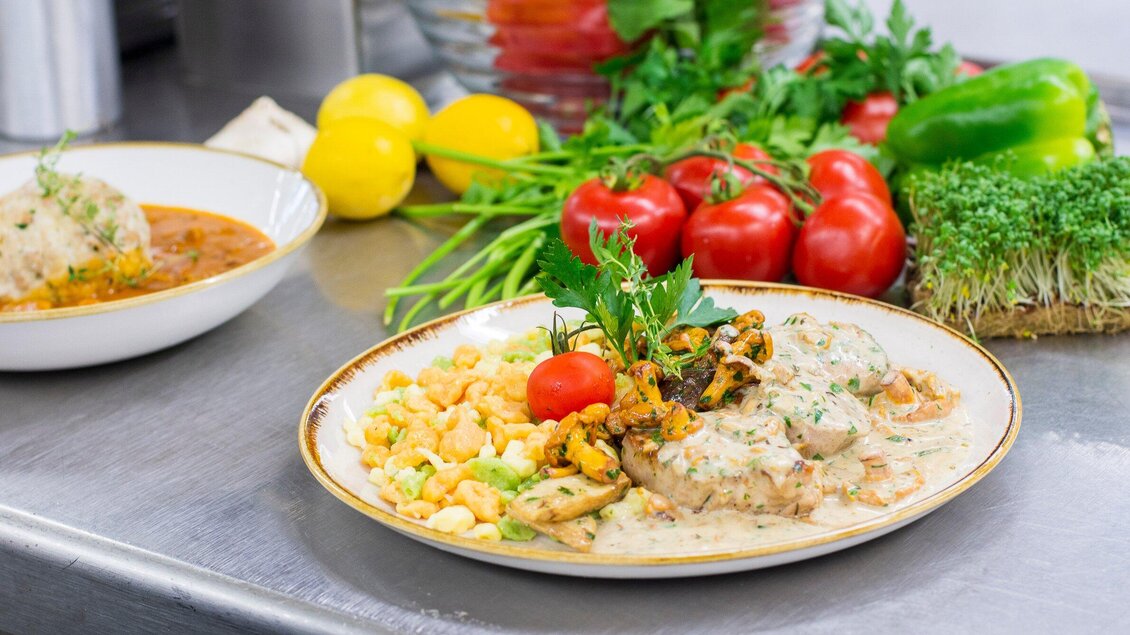 Ein Teller mit schmackhaftem Essen, darunter Fleisch, Gemüse und eine kreative Soße. Im Hintergrund sind frisches Gemüse und Kräuter sichtbar. | © Urdlwirt Hotel - Restaurant
