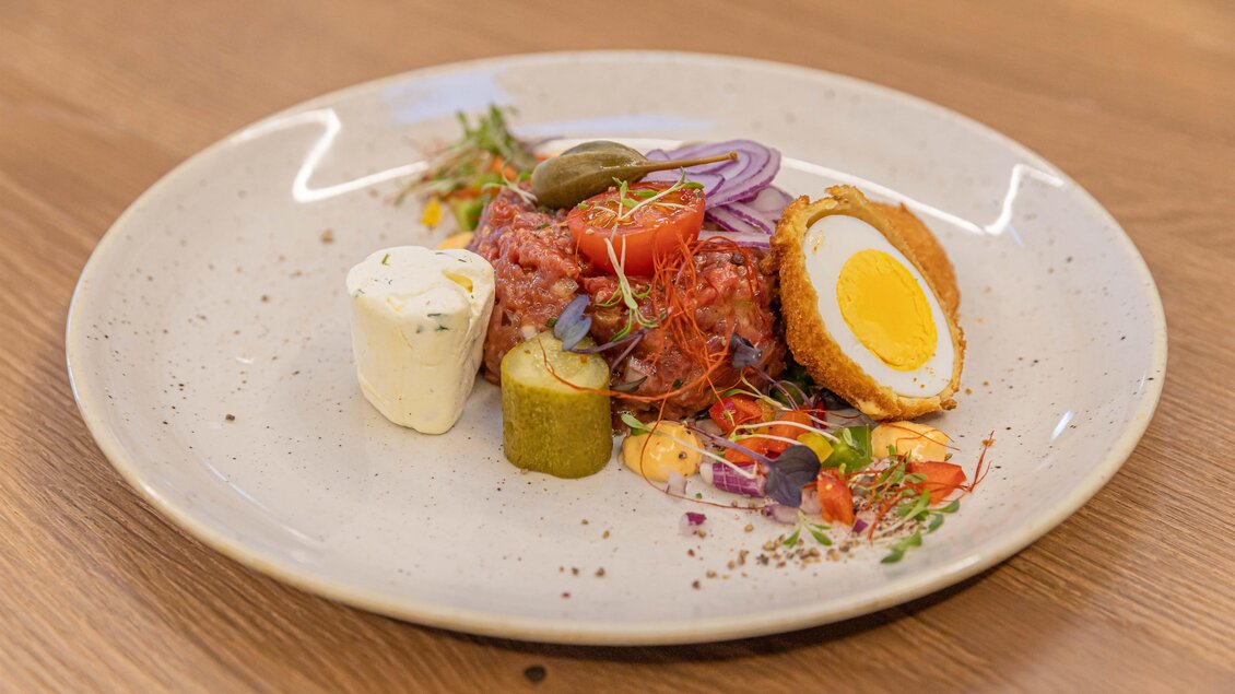 Ein schön angerichteter Teller mit Hackfleisch, einem belegten Ei, sowie verschiedenen Gemüse- und Kräuterbeigaben. Die Präsentation ist appetitlich und farbenfroh. | © Urdlwirt Hotel -  Restaurant