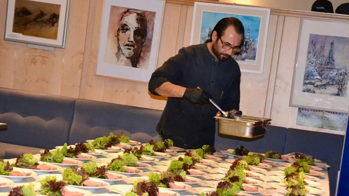 Ein Koch bereitet mehrere Teller mit Salat an einem langen Tisch vor. Im Hintergrund sind Kunstwerke an der Wand zu sehen. | © VAZ Niklasdorf