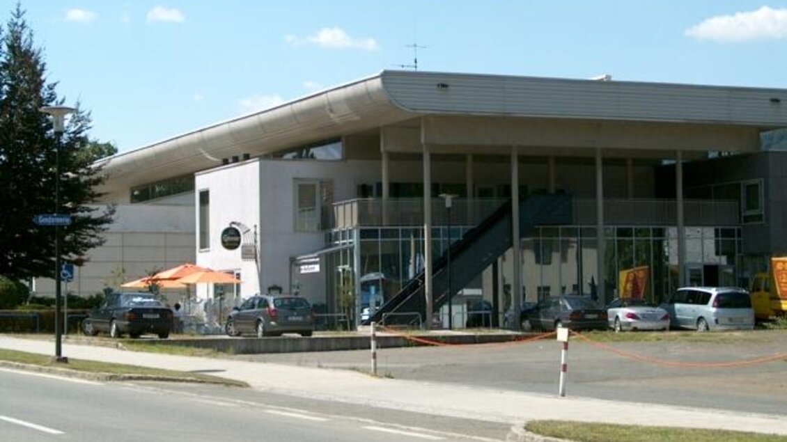Ein modernes Gebäude mit großen Fenstern und einem begrünten Parkplatz. Im Vordergrund sind parkende Autos und ein Gehweg zu sehen. | © VAZ Niklasdorf