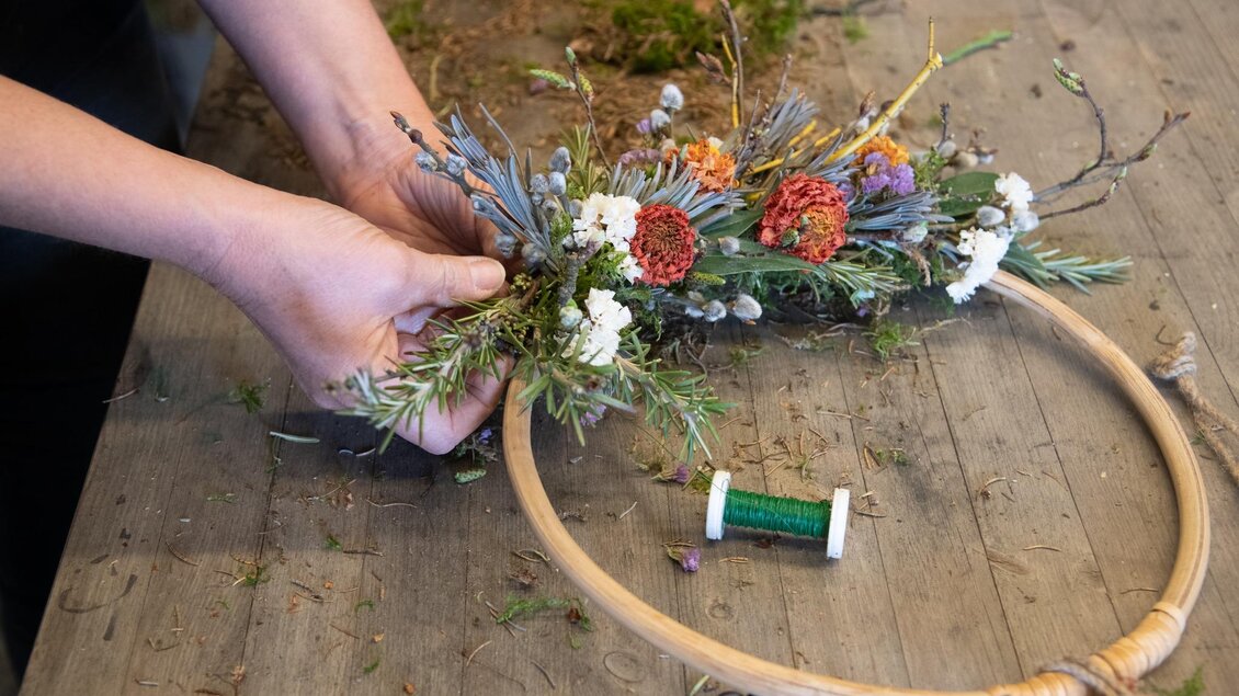 Eine Person gestaltet einen Blumenkranz aus verschiedenen Pflanzen und Trockenblumen. Im Hintergrund ist ein Holztisch mit weiteren Materialien zu sehen. | © Vom Hügel