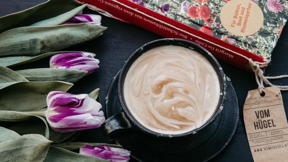 Ein Buch über biologische Blumen liegt auf einem Tisch. Daneben stehen lila Tulpen und eine Tasse mit Kaffee. | © Vom Hügel