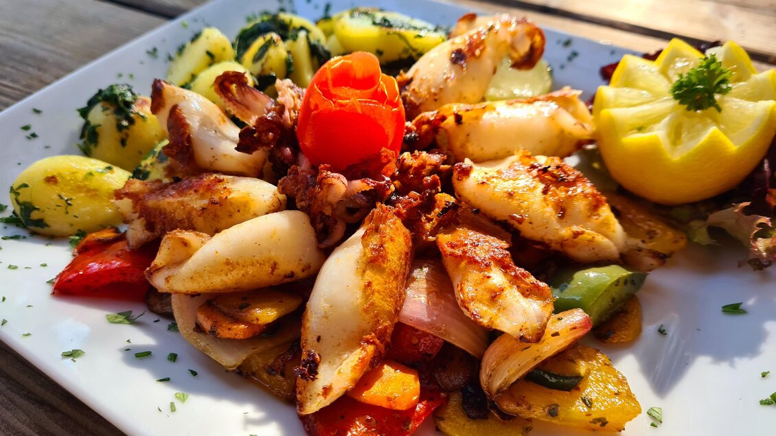 Ein schön angerichteter Teller mit gegrilltem Oktopus, buntem Gemüse und Kürbisscheiben. Dazu gibt es eine Zitrone und eine rote Blume als Dekoration. | © Platzhirsch-Valek