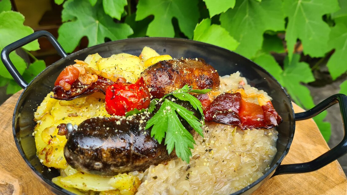 Ein rustikales Gericht in einer Pfanne, bestehend aus Würstchen, Kartoffeln und Gemüse. Das Essen ist mit frischer Petersilie garniert und vor einem grünen Hintergrund angerichtet. | © Platzhirsch-Valek