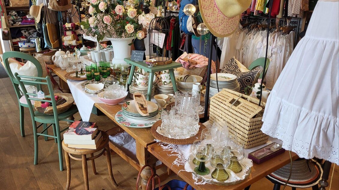 Ein gemütlicher Vintage-Laden mit einem Tisch voller antiker Gläser, Geschirr und Dekorationsartikeln. Im Hintergrund sind bunte Kleidung und Blumenarrangements zu sehen. | © Petra Kirchschlager