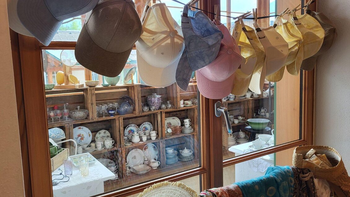 Eine Reihe von bunten Baseballmützen hängt an einer Wäscheleine vor einem Fenster. Im Hintergrund sind verschiedene Geschirrteile in Regalen sichtbar. | © Petra Kirchschlager