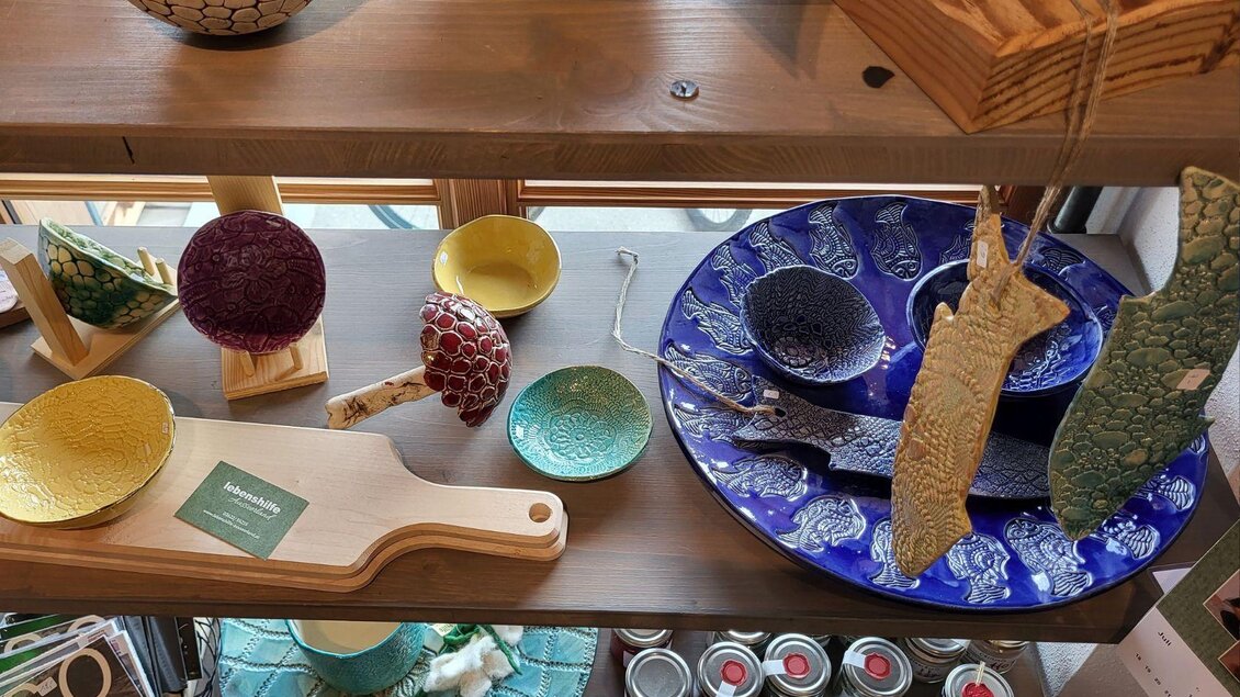 Bunte Keramikschalen und Teller auf einem Holzregal. Eine hölzerne Servierplatte und verschiedene dekorative Elemente sind ebenfalls zu sehen. | © Petra Kirchschlager