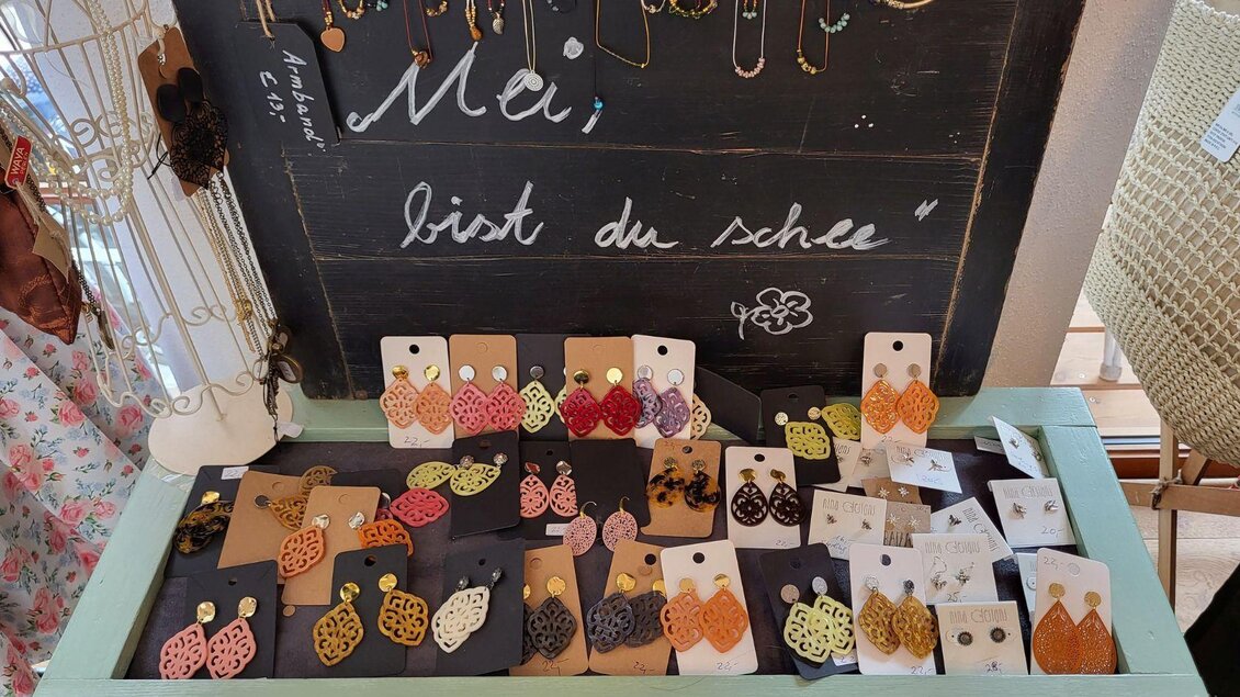 Bunte Ohrringe in verschiedenen Designs und Farben sind in einer schönen Auslage präsentiert. Im Hintergrund steht der Schriftzug "Mei, bist du schee". | © Petra Kirchschlager