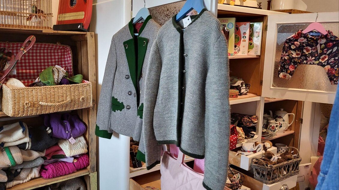 Zwei graue Strickjacken hängen an Kleiderbügeln in einem Geschäft. Im Hintergrund sind verschiedene handgefertigte Produkte und farbige Stoffe zu sehen. | © Petra Kirchschlager
