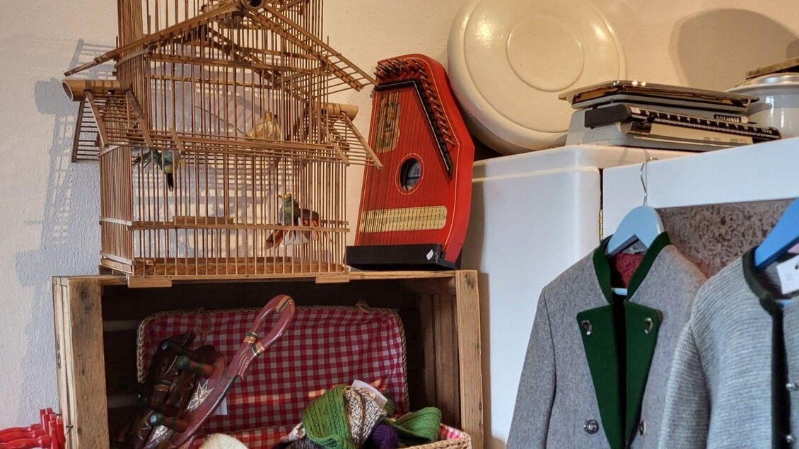 Ein gemütlicher Raum mit einer dekorativen Vogelvoliere und einem alten Musikinstrument. Darunter befindet sich eine Kiste mit bunten Gegenständen und zwei aufgehängten Kleidungsstücken. | © Petra Kirchschlager