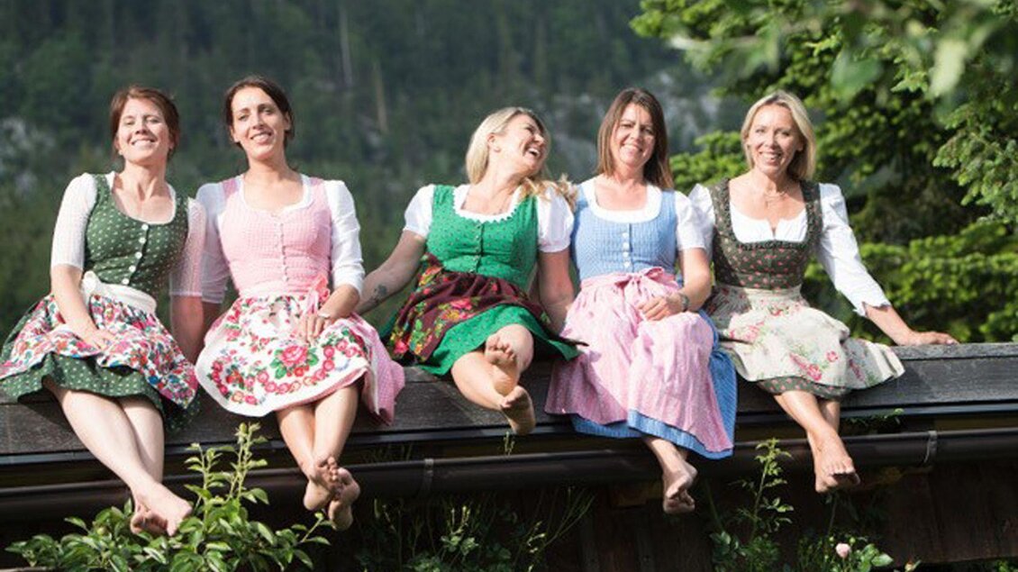 Fünf Frauen in traditionellen Trachten sitzen auf einem Dach. Sie lächeln und genießen den sonnigen Tag in einer schönen Umgebung. | © Coco Kammerer