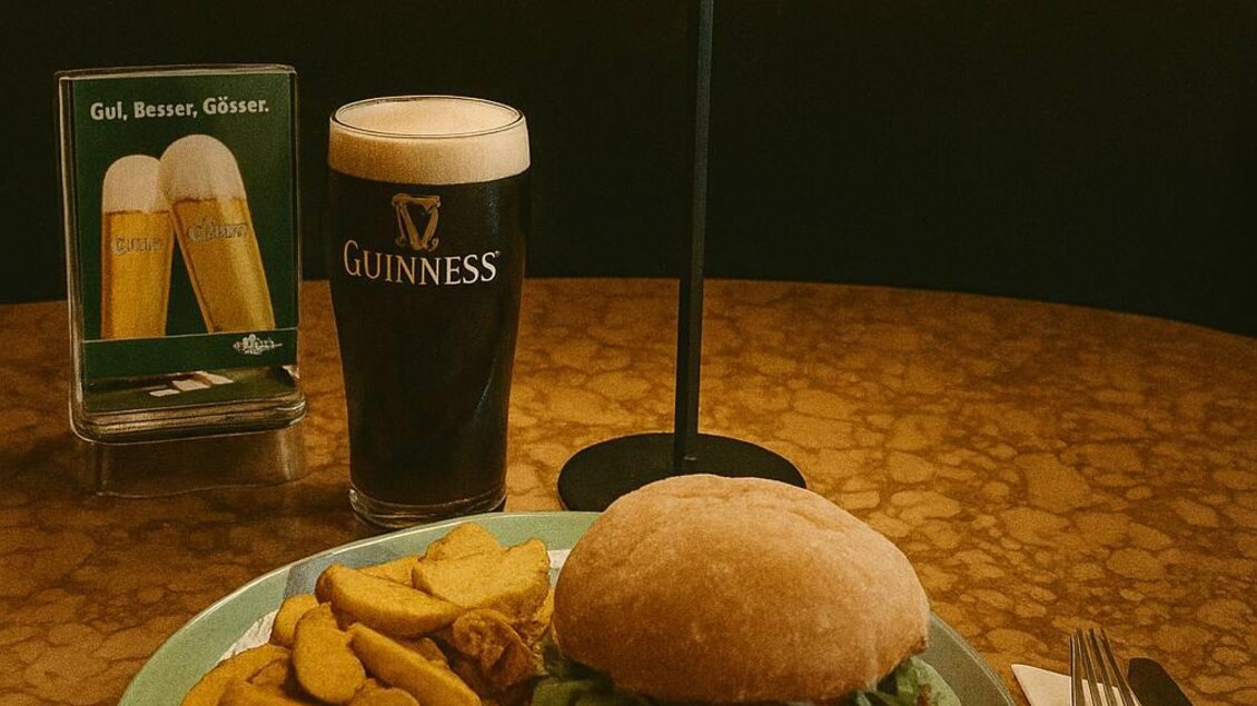 Ein saftiger Burger mit Pommes und Rotkohl auf einem Teller. Dazu ein Glas Guinness auf einem Tisch mit einer kleinen Lampe im Hintergrund. | © Victor Muntean