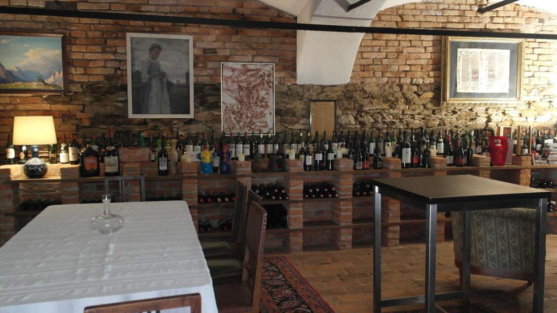 Ein gemütlicher Weinkeller mit Holzregalen voller Flaschen. An den Wänden hängen Bilder und eine Tischgruppe lädt zum Verweilen ein. | © Vinariat Maier