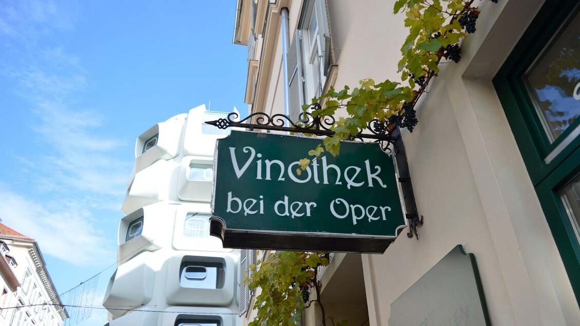 Ein Schild mit der Aufschrift "Vinothek bei der Oper" hängt an einem Gebäude. Im Hintergrund sind moderne Architekturelemente und Weinranken zu sehen. | © Vinothek bei der Oper