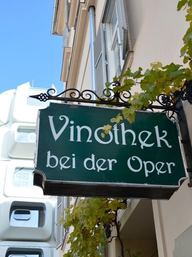 Ein Schild mit der Aufschrift "Vinothek bei der Oper" hängt an einem Gebäude. Im Hintergrund sind moderne Architekturelemente und Weinranken zu sehen. | © Vinothek bei der Oper
