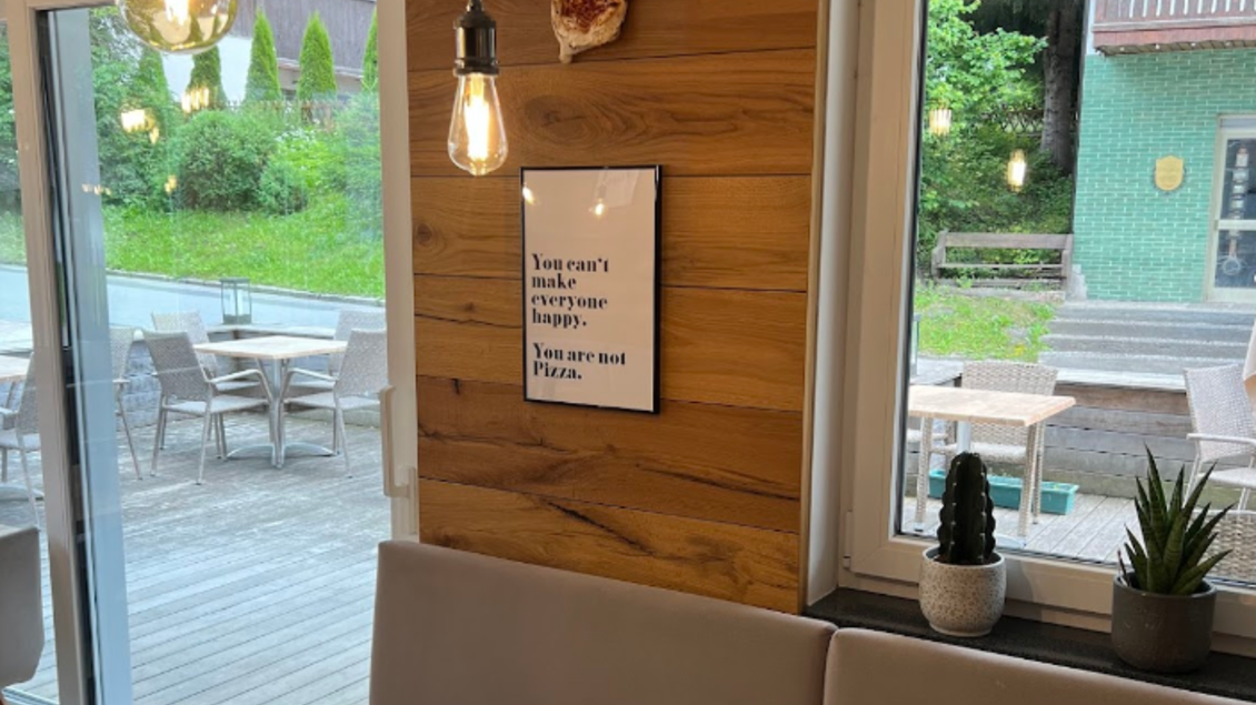 Ein gemütliches Café mit einer holzverkleideten Wand und großen Fenstern. An der Wand hängt ein inspirierendes Zitat und es gibt einige Pflanzen auf der Fensterbank. | © Vivo Pizzeria